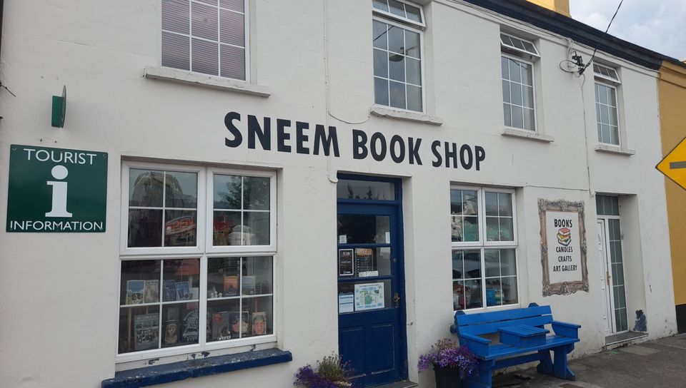 The white two-storey frontage of Sneem Book Shop with SNEEM BOOK SHOP in black lettering, a navy front door, flower pots, a blue bench and the green Tourist Information sign