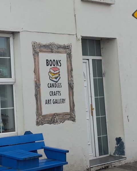 A hand-painted picture frame on the white wall of Sneem Book Shop reading Books, Candles, Crafts, Art Gallery, above the blue bench outside the shop